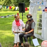 Zwei Personen sitzen auf einer kleinen Bühne im Park und lesen gemeinsam aus einem Buch vor Publikum.