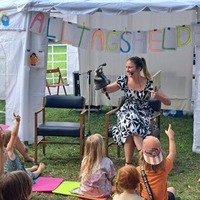 Eine Frau in einem schwarz-weißen Kleid sitzt auf einem Stuhl und hält eine Puppe in der Hand. Sie liest vor einer Gruppe von Kindern, die aufmerksam zuhören und auf sie zeigen. Im Hintergrund ist ein Zelt mit dem Banner 'Alltagsheld' zu sehen.