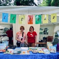 Zwei Frauen stehen hinter einem Tisch mit einer Auswahl an Kinderbüchern. Über dem Tisch hängt ein bunter Banner mit der Aufschrift 'Lesebar'. Die Frauen lächeln und begrüßen Besucher.