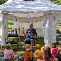 Person liest unter einem weißen Pavillon aus einem Buch vor, mehrere Kinder sitzen draußen auf dem Boden und hören zu.